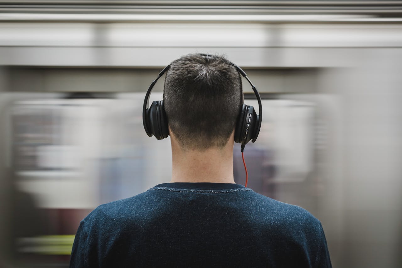Rear view of a man with headphones standing at a subway platform, blurred train passing by.