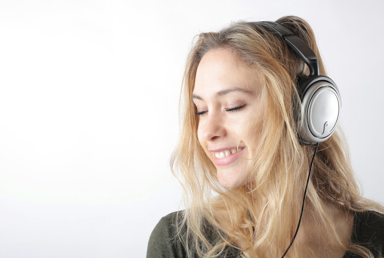 A cheerful woman with blonde hair listens to music on headphones, eyes closed, on a white background.