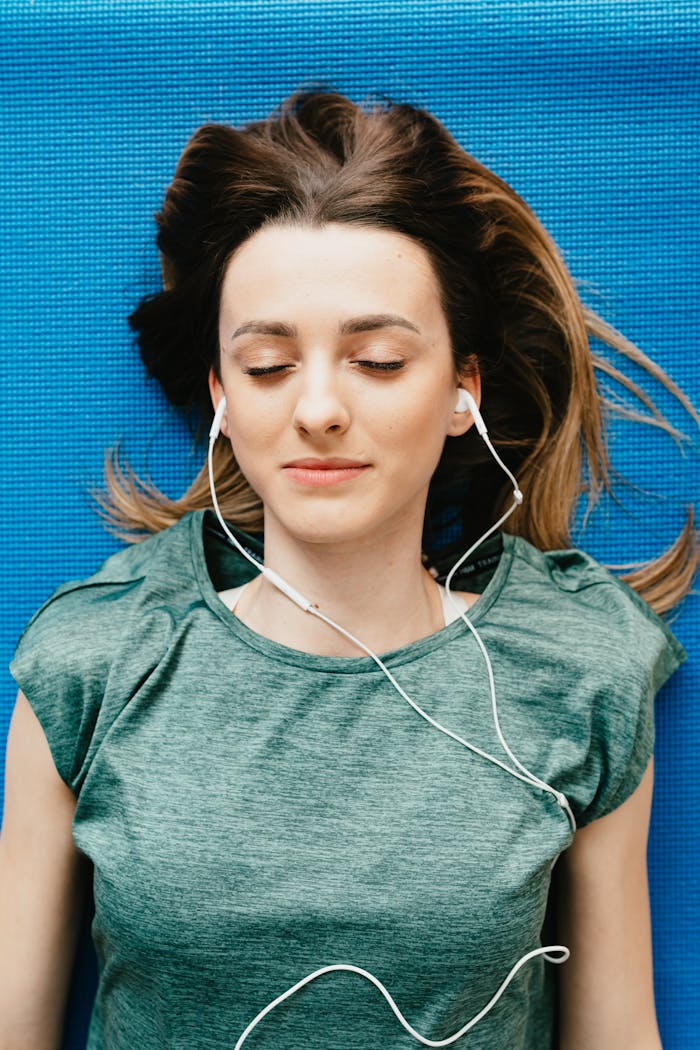 From above of slim peaceful lady in casual clothes and earphones resting on yoga mat with closed eyes during meditation at home