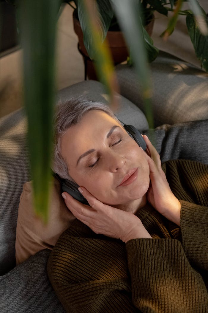 A mature woman with gray hair relaxes on a sofa, enjoying music through wireless headphones.