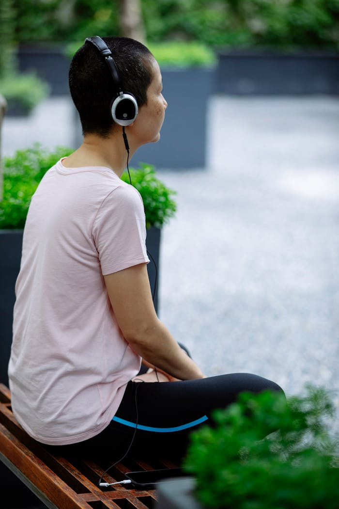 Faceless woman in casual clothes sitting with crossed legs on bench with phone in park near green plants in pots with headphones in summer day